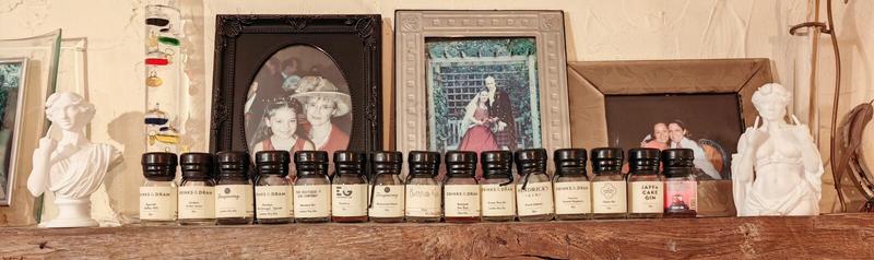 Photo: Fifteen empty small glass bottles of gin arranged in a row on a dark wooden surface, flanked by white decorative busts and framed photographs. From left to right, the bottles read: "DRINKS BY THE DRAM Sipsmith Raffles 1915 Gin", "DRINKS BY THE DRAM Gordon's Sicilian Lemon Gin", "Tanqueray London Dry Gin", "DRINKS BY THE DRAM Darnley's Navy Strength - Spiced London Dry Gin", "THAT BOUTIQUE-Y GIN COMPANY Moonshot Gin", "EDINBURGH GIN Raspberry Gin", "Tanqueray Blackcurrant Royale Gin", "Bathtub Gin", "DRINKS BY THE DRAM Boatyard Sloe Boat Sloe Gin", "HENDRICK'S GIN Grand Cabaret Gin", "DRINKS BY THE DRAM Greater Than Gin", "DRINKS BY THE DRAM Caorunn Scottish Raspberry Gin", "THE LAKES DISTILLERY Classic Gin", "JAFFA CAKE GIN", and what appears to be "THAT BOUTIQUE-Y GIN COMPANY Cherry Gin" with a pink label. Each bottle has a black screw cap. Behind the bottles are four framed photographs: the ornate dark wooden oval frame containing the 25-year-old photograph of an 8-year-old girl and woman in red, a light-coloured rectangular frame showing two people in formal dress in a garden, a brown wooden frame with another photograph, and additional family photos. On the far left is a tall decorative bottle with coloured horizontal stripes. Two white classical-style busts bookend the display. The scene is set against a cream or beige textured wall.Claude is AI and can make mistakes. Please double-check responses.