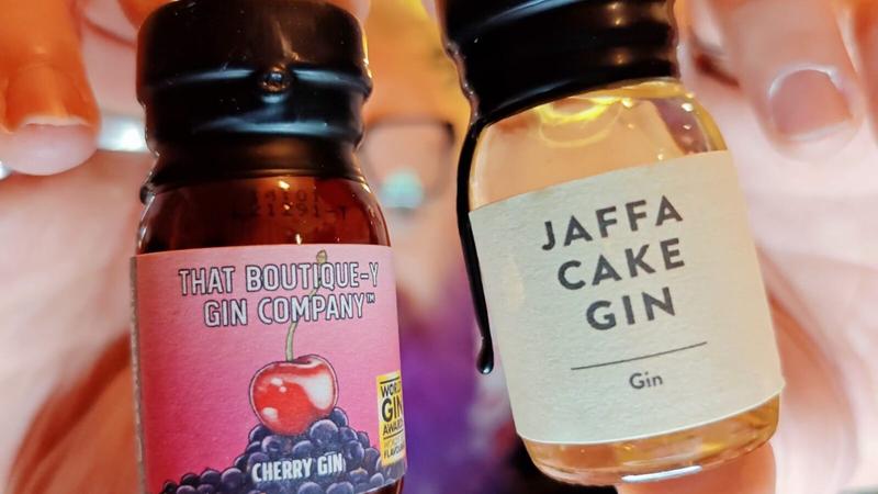Photo: Two hands holding small glass bottles of gin against a soft, warm peachy-pink blurred background. On the left, a bottle with a black screw cap containing dark reddish-brown liquid. The label features a pink and purple gradient background with "THAT BOUTIQUE-Y GIN COMPANY" text at the top, an illustration of a red cherry and dark berries below, and "CHERRY GIN" text. A small yellow badge showing "WORLD GIN AWARDS" is visible on the label. On the right, a bottle with a black screw cap containing pale golden liquid. The label is cream-coloured with simple black text reading "JAFFA CAKE GIN" followed by a horizontal line and "Gin" below. The background shows warm lighting and indistinct soft shapes suggesting an interior setting.
