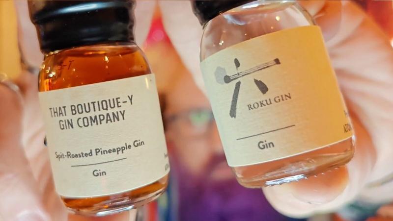 Photo: Two hands holding small glass bottles of gin against a soft, warm blurred background. On the left, a bottle with a black screw cap containing amber or golden-brown liquid. The cream-coloured label reads "THAT BOUTIQUE-Y GIN COMPANY" at the top, followed by "Spit-Roasted Pineapple Gin" and "Gin" below. On the right, a slightly larger bottle with a black screw cap containing pale golden liquid. The cream-coloured label features a simple illustration of a Japanese-style design or character at the top, with "ROKU GIN" text and "Gin" below. The background shows soft focus with warm peachy tones and hints of purple, suggesting people and warm interior lighting.