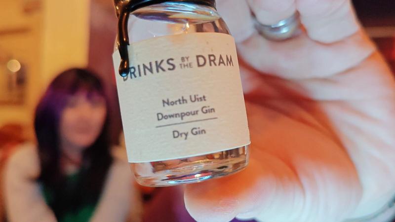 Photo: A hand holding a small glass bottle of gin in sharp focus towards the camera against a warm, softly blurred background. The bottle has a black screw cap and a cream-coloured label reading "DRINKS BY THE DRAM" at the top, followed by "North Uist Downpour Gin", a horizontal line, and "Dry Gin" below. The bottle contains clear liquid. The background is deliberately out of focus, showing warm peachy-orange and purple tones with indistinct blurred shapes suggesting people and interior furnishings. Light bokeh is visible in the upper left corner.