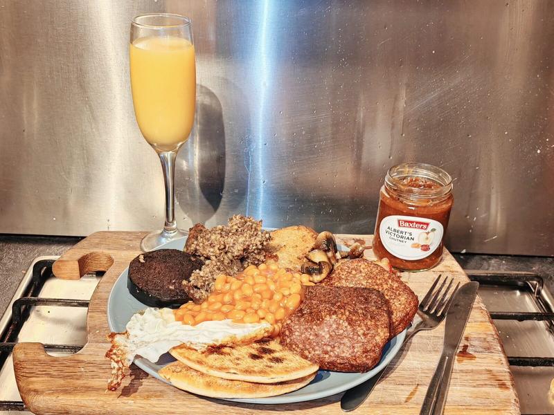Photo: A full Scottish breakfast on a white plate with a champagne flute of orange juice and a jar of Baxters Alberta Victorian Pickle. The plate contains black pudding, white pudding, haggis slices, baked beans, a fried egg, two grilled potato scones, Lorne sausage, and bacon, all arranged against a stainless steel splashback.