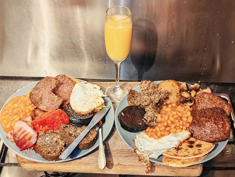 Photo: Two full Scottish breakfast plates side by side on a wooden board, each with a champagne flute of orange juice between them. Both plates are loaded with the full works: black pudding, white pudding, haggis, beans, eggs, potato scones, sausage, bacon, and grilled tomato. Because one breakfast is never enough.