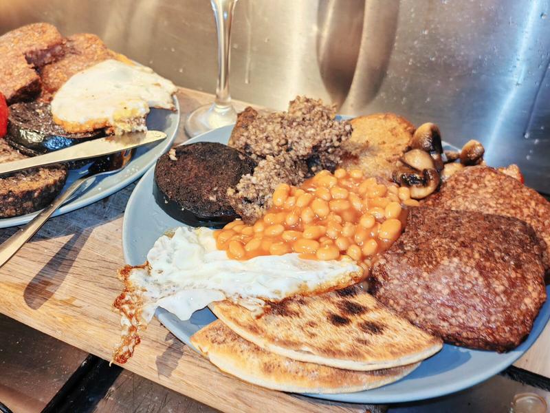 Photo: Close-up of a Scottish breakfast plate showing the layered construction: fried egg on black pudding, with haggis slices, beans spilling across the plate, potato scones, Lorne sausage, and bacon. The champagne flute stands guard in the background.