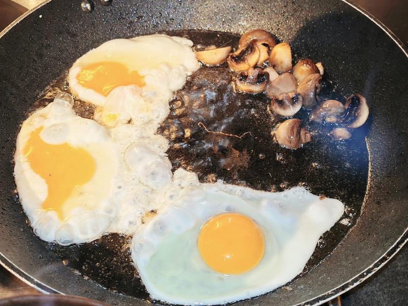 hoto: Three fried eggs and sliced mushrooms sizzling in a black frying pan. The eggs have vibrant orange yolks and are at that perfect stage where the whites are just set. This is where the magic happens.