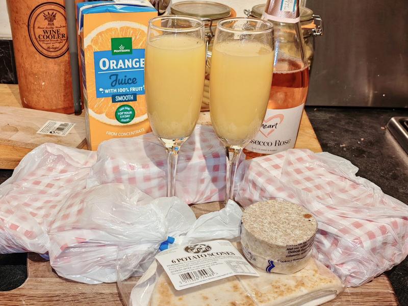 Photo: Two champagne flutes filled with orange juice standing between plastic-wrapped ingredients on gingham tea towels. A bottle of Prosecco Rosé lurks behind them. Breakfast labels visible on the potato scones. We're having Buck's Fizz, obviously.