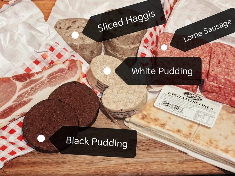 Raw ingredients laid out on a wooden chopping board: sliced haggis, white pudding rounds stacked up, black pudding slices, bacon rashers on gingham paper, Lorne sausage squares, and potato scones in packaging. Everything present and correct for inspection.