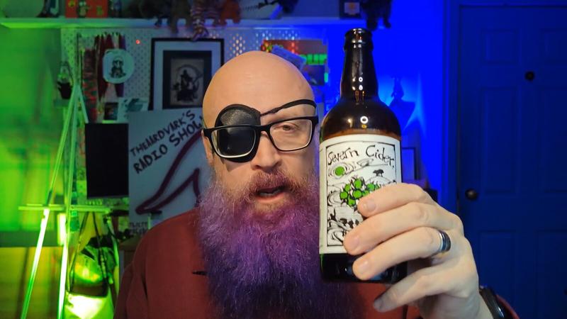 Photo: theaardvark holding a bottle of Severn Cider towards the camera. He's wearing black-framed glasses over an eye patch covering his right eye, giving a pirate aesthetic. His mouth is open mid-speech. The bottle has a cream-coloured label with green apple illustrations and "Severn Cider" text. Green and blue ambient lighting illuminates the background pegboard wall with the TTHEAARDVARK’S RADIO SHOW. Silver ring visible on his hand holding the bottle.