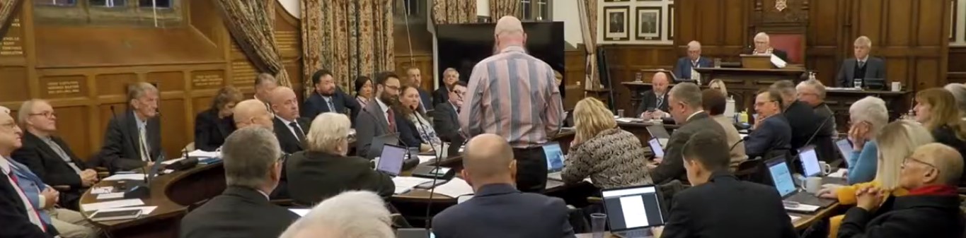 Photo of a formal council chamber with dark wood panelling and ornate decor. Around 30 people are seated at a large curved table, most in suits, with laptops and papers in front of them. A bald man in a blue and white striped shirt stands with his back to the camera, addressing the room — standing out somewhat from the sea of formal attire. At the far end, several officials are seated at a raised platform. Everyone appears to be paying attention, which makes a nice change.