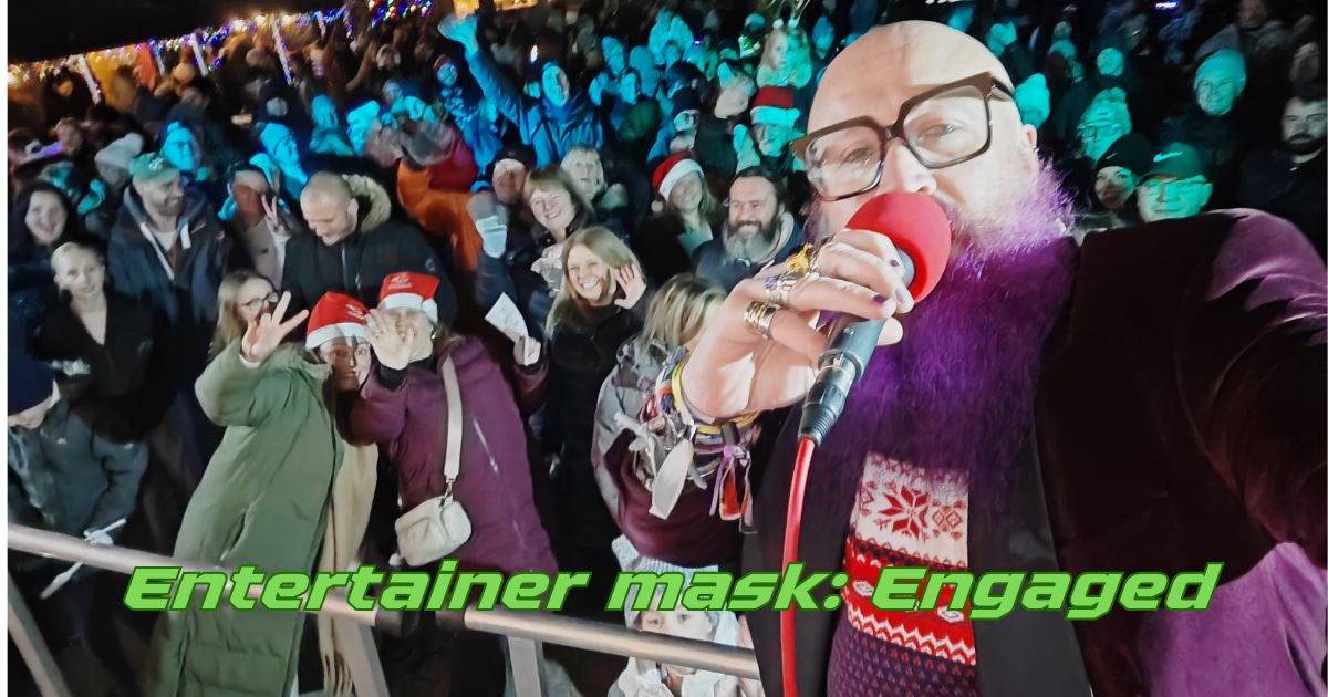 A bald man with a purple beard, and glasses holds a microphone with large red foam cover in front of a large, happy crowd at a nighttime outdoor event. He is wearing a Christmas jumper and appears to be having an unreasonable amount of fun. Text overlaid on the image reads "Entertainer mask: Engaged" in green robot font. The mask is, by all available evidence, working.