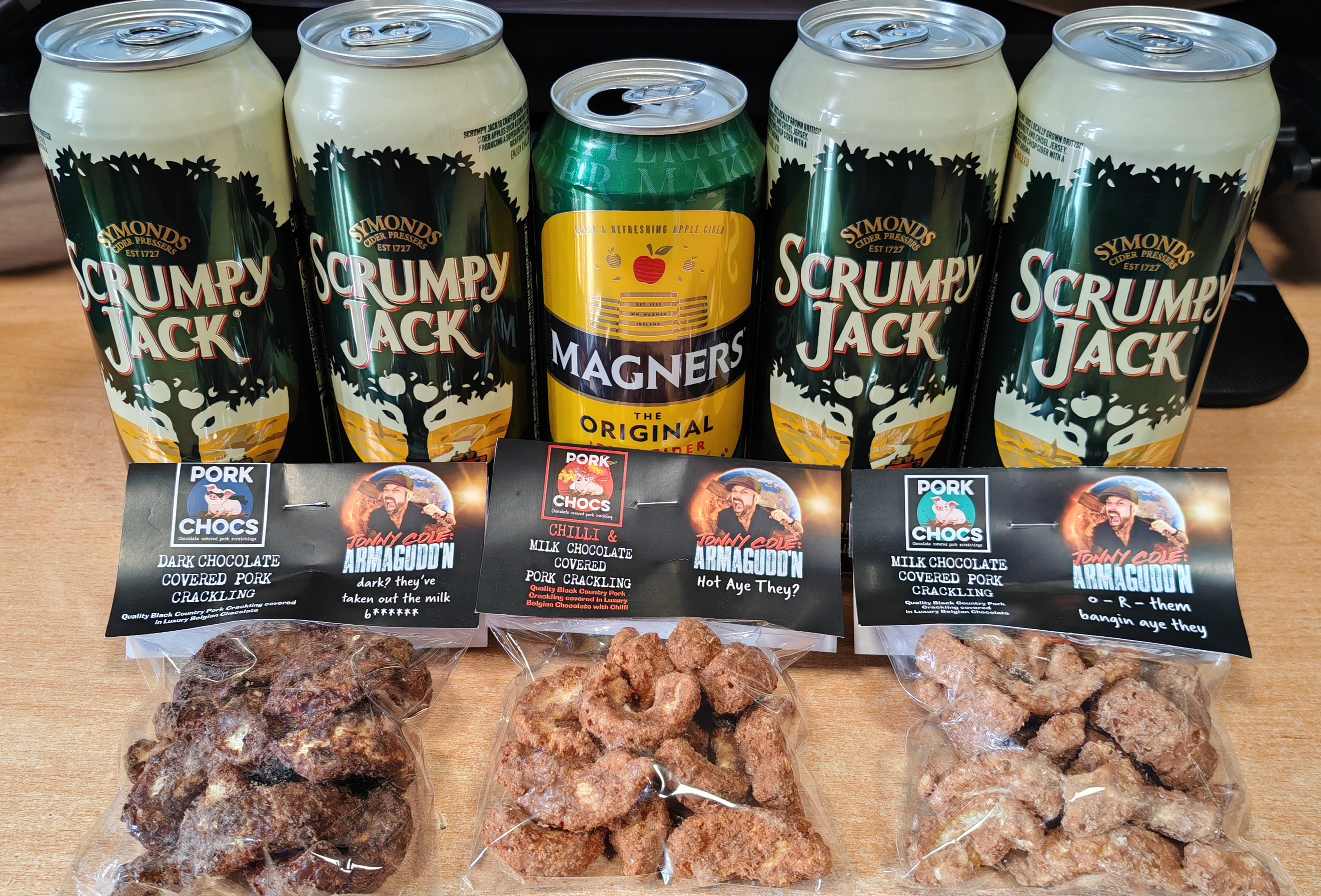 Photograph of five cans of cider arranged in a row — four Symonds Scrumpy Jack and one Magners Original Apple Cider in the centre — with three clear bags of Pork Chocs chocolate-covered pork crackling in front of them. The Pork Chocs bags are branded with the "Jonny Cole: Armagudd'n" label and come in three varieties: Dark Chocolate Covered Pork Crackling ("dark? they've taken out the milk"), Chilli & Milk Chocolate Covered Pork Crackling ("Hot Aye They?"), and Milk Chocolate Covered Pork Crackling ("o-R-them bangin aye they"). Everything is arranged on a wooden surface. The crackling looks exactly as alarming as it sounds.
