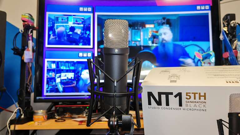 A Røde NT1 5th Generation Black studio condenser microphone, in its shock mount, posed next to its own packaging like it's about to sign a record deal. Behind it, a large monitor displays a multi-camera stream layout with purple and blue lighting. The studio is visibly lived-in.