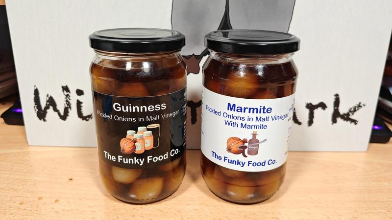 Two sealed jars of Funky Food Co. pickled onions on a wooden surface against a white backdrop with partial text visible. Left jar has a black label for "Guinness Pickled Onions in Malt Vinegar" with illustrations of onions and Guinness bottles. Right jar has a white label for "Marmite Pickled Onions in Malt Vinegar With Marmite" with similar illustrations including a Marmite jar.