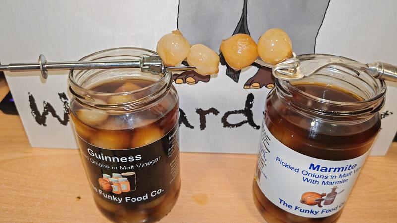 Photo: Both jars with metal forks holding pickled onions above them. Two onions from each jar rest on the fork prongs, showing the colour difference - the Guinness onions appearing lighter and more golden, the Marmite onions showing a slightly darker, more amber tone. The white backdrop features partial lettering, and the composition demonstrates the visual comparison between the two varieties.