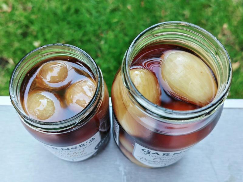 Photo: Top-down view into both opened Garner's jars showing the pickled onions floating in their dark amber malt vinegar. The left jar contains smaller shallots whilst the right jar shows larger original pickled onions. The glass jar threads are visible, and the rich colour of the malt vinegar is clearly displayed against the green grass background.