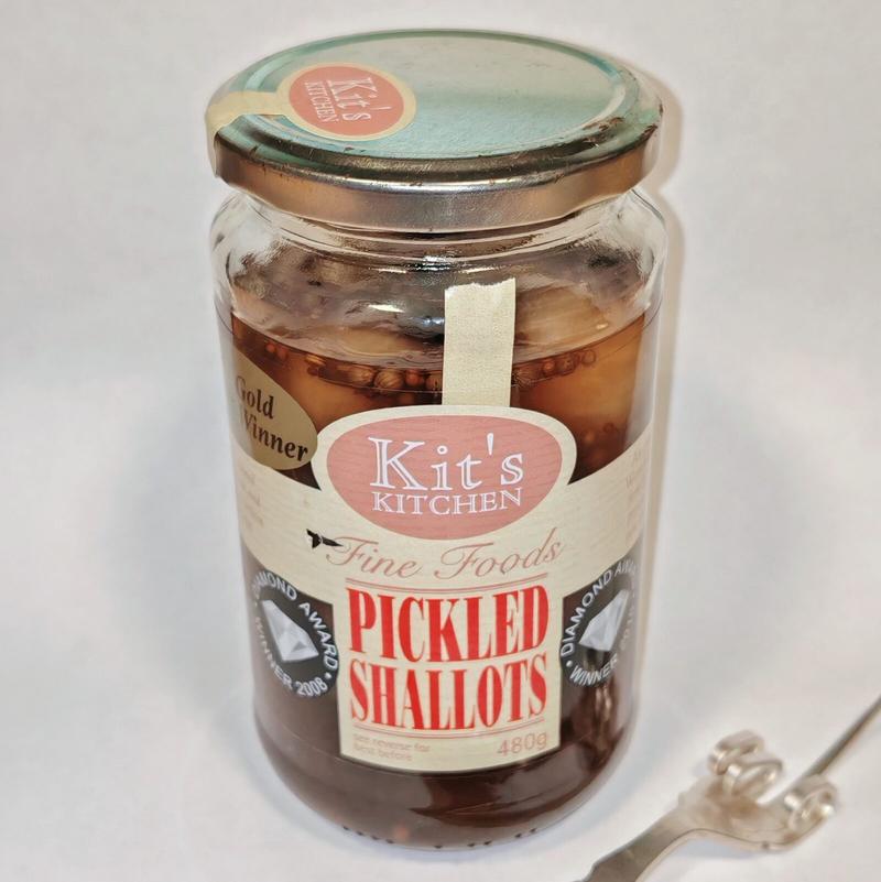 Photo: Sealed jar of Kit's Kitchen Fine Foods Pickled Shallots (480g) with a copper-coloured lid and cream-coloured label featuring the brand's logo and "Gold Award Winner" badges. The jar shows the pickled shallots and spices clearly visible through the glass, with a metal fork partially visible at the bottom right of the frame against a white background. Those award badges suggest someone, somewhere, takes their pickled shallots very seriously indeed.RetryClaude can make mistakes. Please double-check responses.