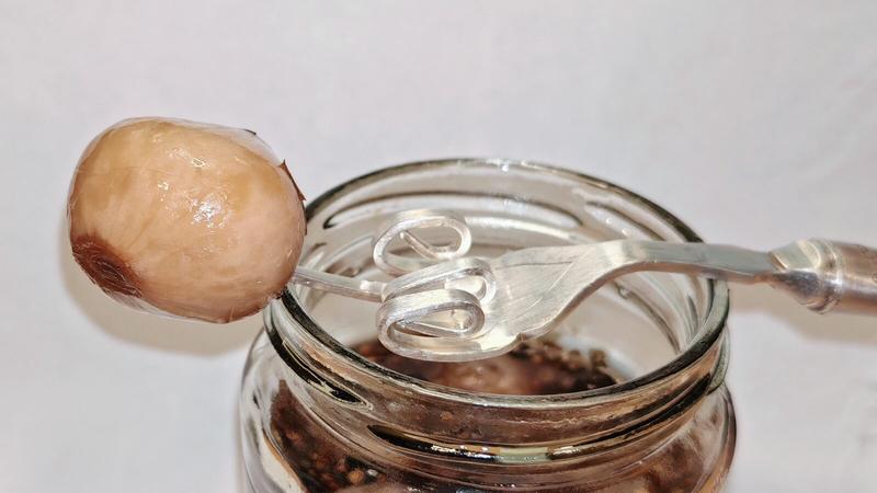 Photo: Single pickled shallot on a metal fork held above an opened jar against a white background. The shallot appears golden and translucent with a darker brown patch where it's been sliced or where the root end was, showing the characteristic appearance of a properly pickled specimen ready for evaluation. It's the moment of truth - will this be brilliant or bin-worthy?