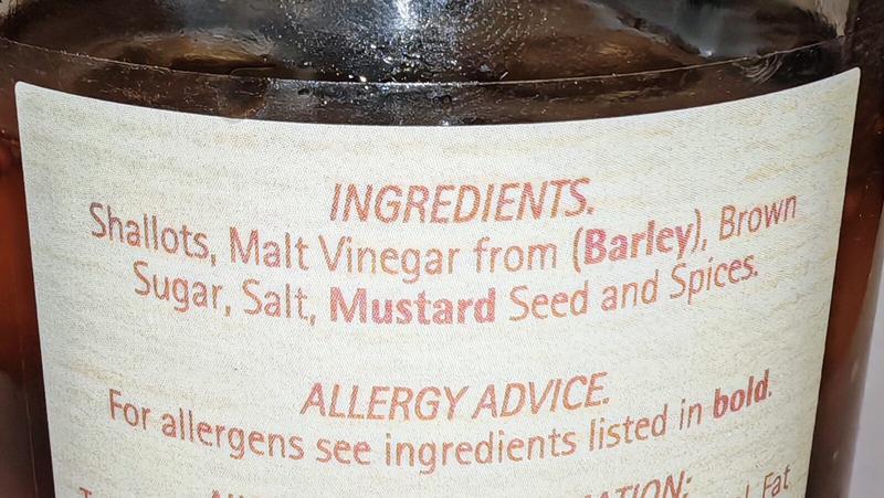 Photo: Close-up of a jar label showing ingredients and allergy information. The label lists "Shallots, Malt Vinegar from (Barley), Brown Sugar, Salt, Mustard Seed and Spices" as ingredients, with allergy advice noting "For allergens see ingredients listed in bold" followed by nutritional information below. A straightforward list that manages to make preserved onions sound almost sophisticated.