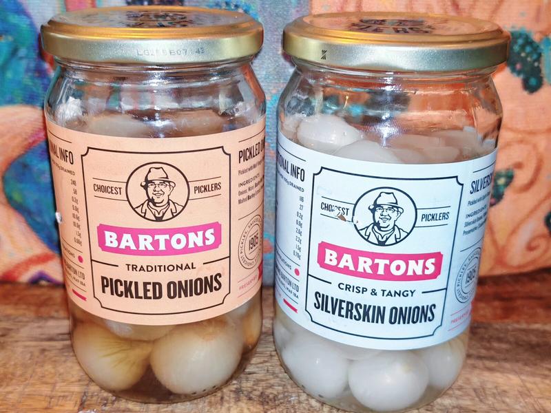 Two glass jars of Bartons pickled onions on a wooden surface. Left jar has a beige label reading "Traditional Pickled Onions" containing pale brown onions. Right jar has a pale blue label reading "Crisp & Tangy Silverskin Onions" containing white onions. Both jars have gold lids and feature the Bartons "Choicest Picklers" logo. Colourful fabric visible in background.