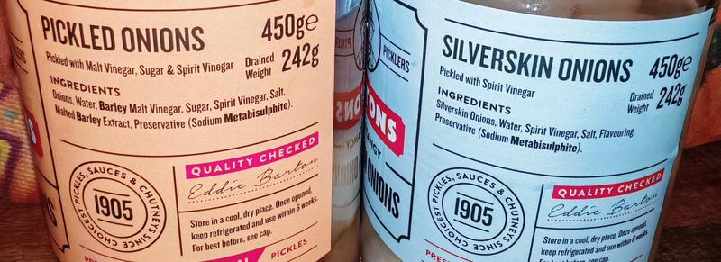 Close-up of jar labels showing ingredients. Traditional Pickled Onions (beige label) lists ingredients: Onions, Water, Barley Malt Vinegar, Sugar, Spirit Vinegar, Salt, Malted Barley Extract, Preservative. Silverskin Onions (pale blue label) lists: Silverskin Onions, Water, Spirit Vinegar, Salt, Flavouring, Preservative. Both show "1905" heritage badge, "Quality Checked" stamp, and Eddie Barton 