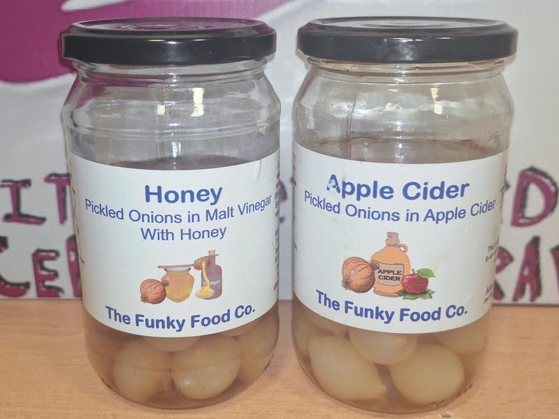 Two sealed jars of The Funky Food Co pickled onions with black lids, showing the full labels. Left: Honey Pickled Onions in Malt Vinegar With Honey. Right: Apple Cider Pickled Onions in Apple Cider. Both jars are approximately half-full of pale pickled onions in clear liquid. Background shows partial purple text.