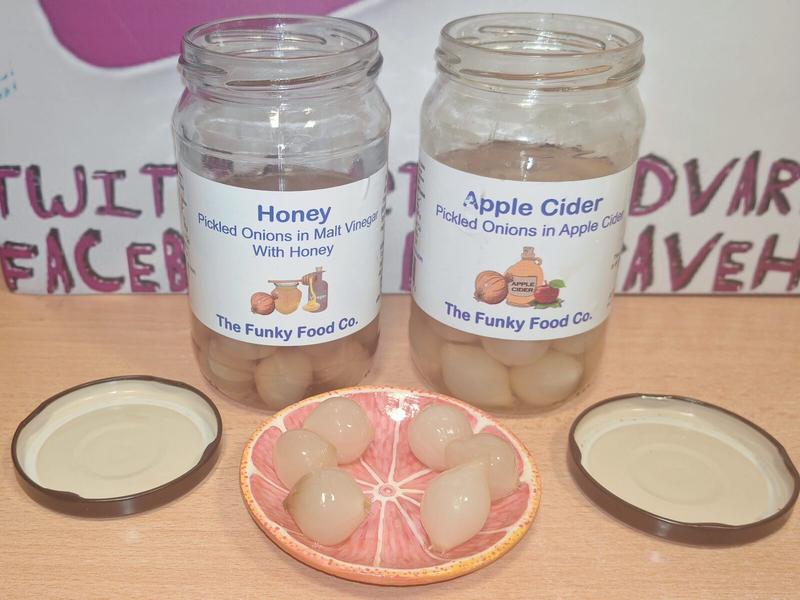 Two opened jars of The Funky Food Co pickled onions on a wooden surface with their lids placed beside them. Left jar: Honey Pickled Onions in Malt Vinegar with Honey. Right jar: Apple Cider Pickled Onions in Apple Cider. In front of the jars is a pink grapefruit-shaped ceramic dish containing five pickled onions arranged in a circle. Background shows partial text giving theaardvark's (former) Twitter (RiP) handle and Facebook address.
