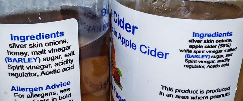 Close-up of ingredient labels on both jars. Left label reads: "Ingredients: silver skin onions, honey, malt vinegar (BARLEY) sugar, salt Spirit vinegar, acidity regulator, Acetic acid. Allergen Advice: For allergens, see ingredients in bold." Right label reads: "Ingredients: silver skin onions, apple cider (50%) white spirit vinegar malted (BARLEY) sugar, salt Spirit vinegar, acidity regulator, Acetic acid. This product is produced in an area where peanuts"