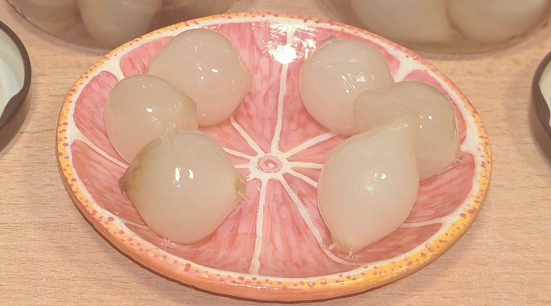 Photo: Ceramic dish shaped and painted as a pink grapefruit slice, viewed from above. Six pale pickled onions are arranged in a circle on the dish's surface, which features detailed segments radiating from a central circle, painted in coral pink with white membrane lines. The rim is painted and textured to resemble grapefruit peel in orange and yellow tones. Liquid from the pickled onions pools in the centre and segments.