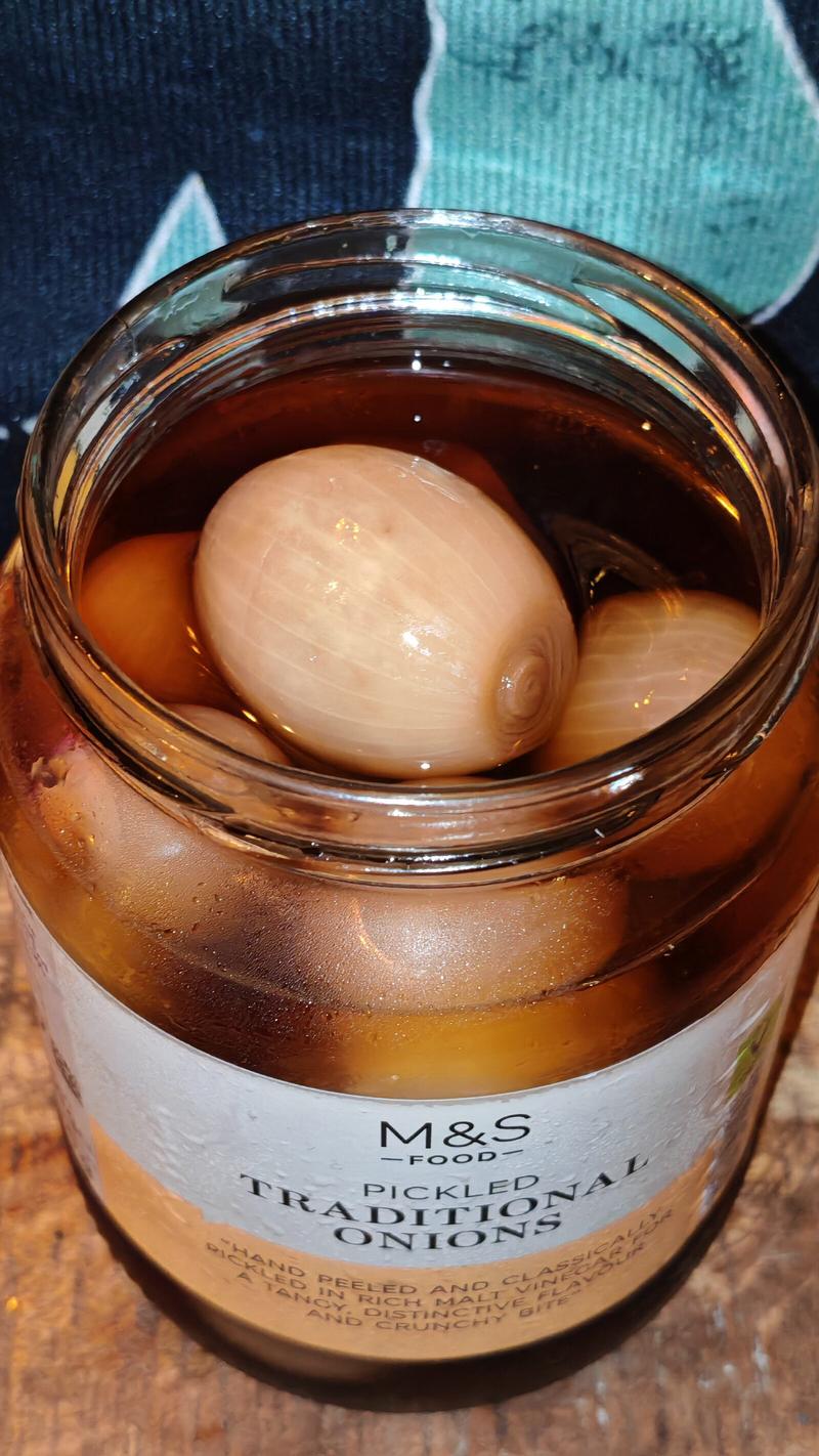 A jar of M&S pickled traditional onions sits open, revealing plump white onions floating in amber vinegar. The jar's label is clearly visible against a colourful knitted background.