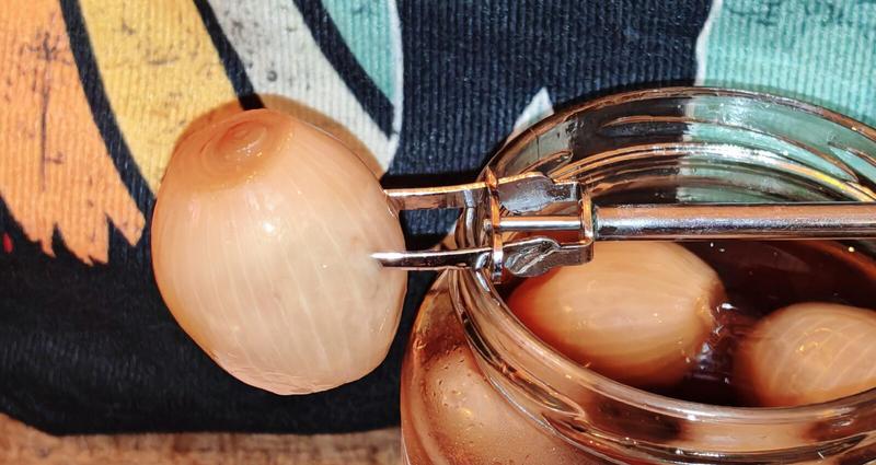 Close-up of silver tongs extracting a perfectly round pickled onion from the jar, with more onions visible floating in the vinegar below. The tongs grip the onion like it's precious cargo.