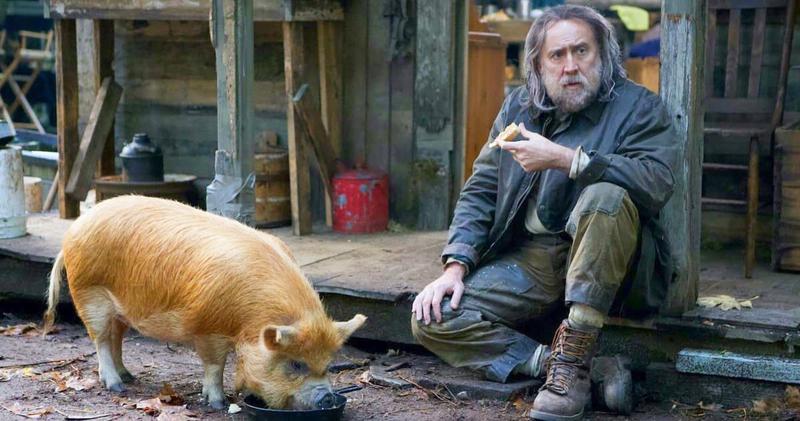 A movie still showing Nicholas Cage looking like a wild-man-from-the-woods stitting on the stoop of a shack with his small, truffle-hunting pig.