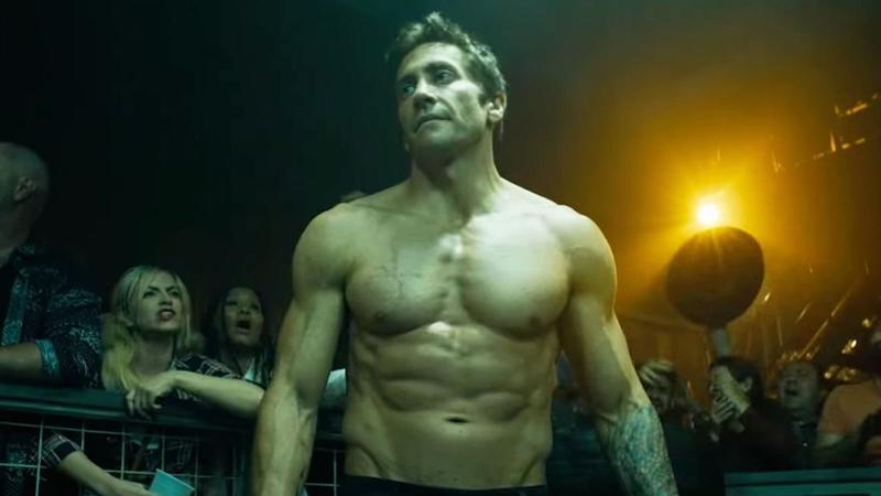 Film still from Road House: a shirtless fighter stands in a crowded, dimly lit fight venue, with spectators leaning on a barrier behind him.